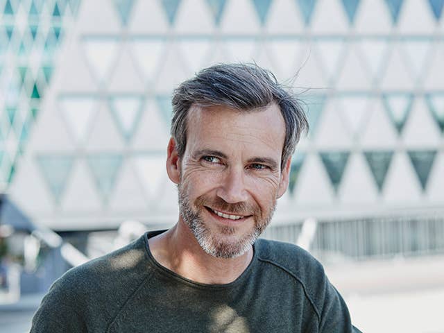 Smiling middle-aged white man with brown/grey hair and a beard stands outdoors in the sunshine