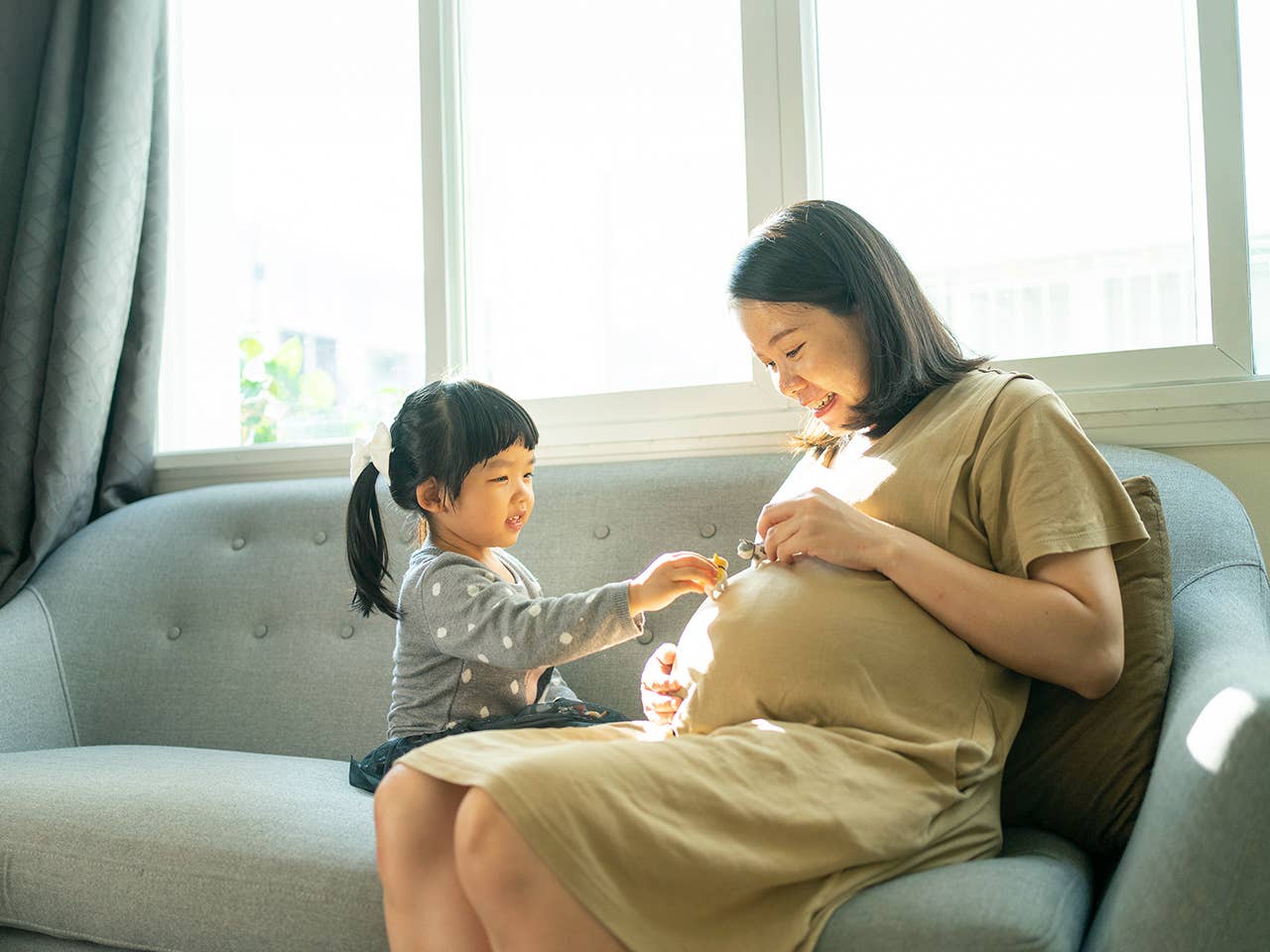 Pregnant Asian woman sits on a couch as her young daughter touches her belly