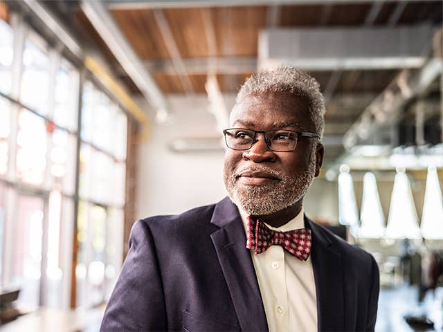 Elderly Black man wearing a navy blue sport coat with a white shirt and bowtie looks out a window