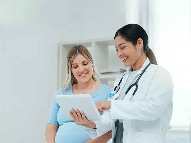 A female doctor is talking to a showing a pregnant female patient something on a tablet