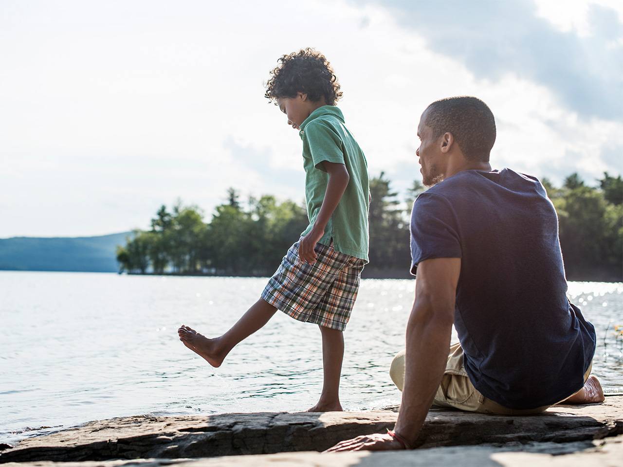 Young Black boy with curly brown hair stands on a large rock near a lake while his father sits next to him