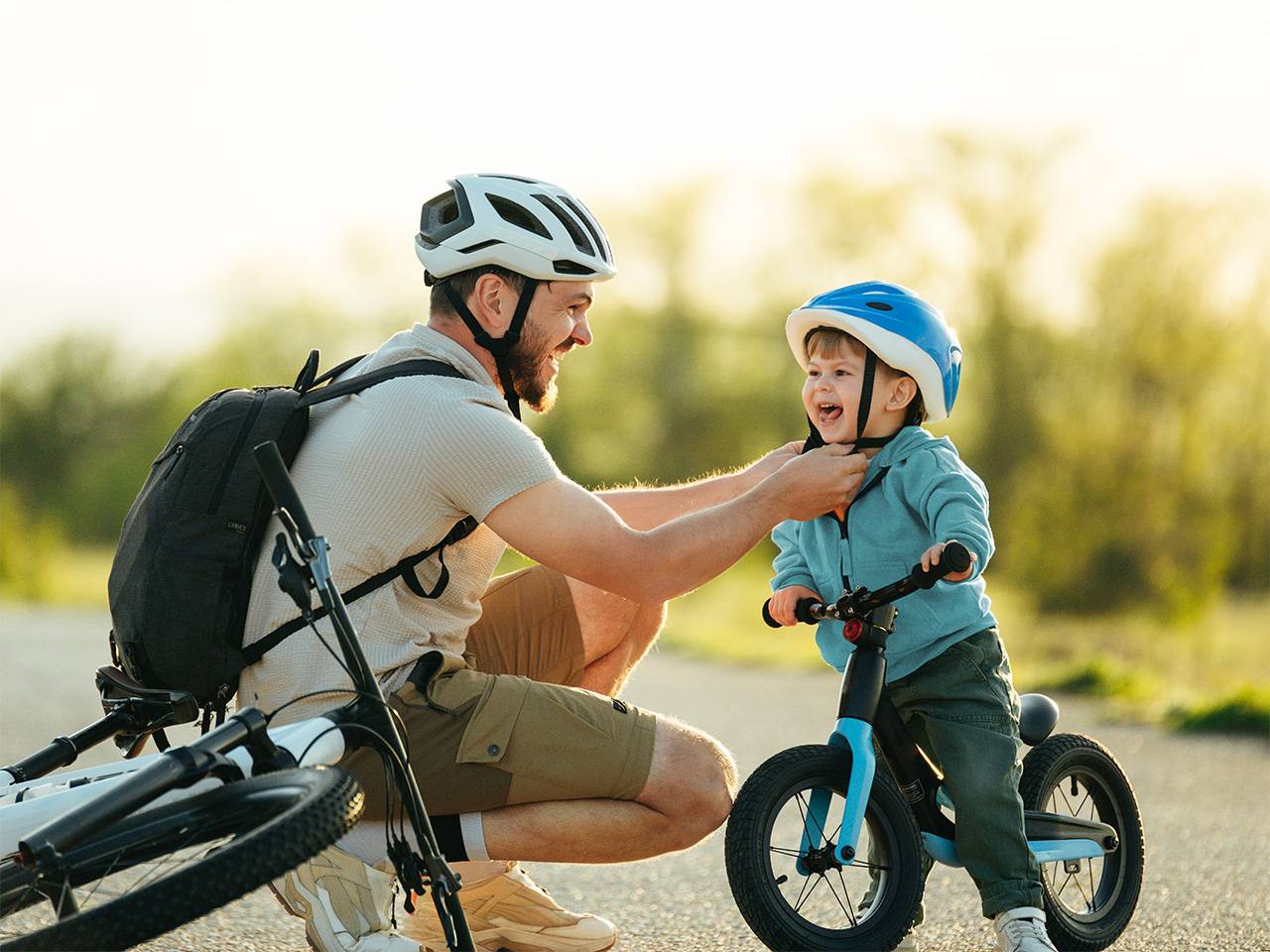 Smiling dad wearing a bike helmet clips a bike helmet on his smiling son while they are outdoors in the sunshine