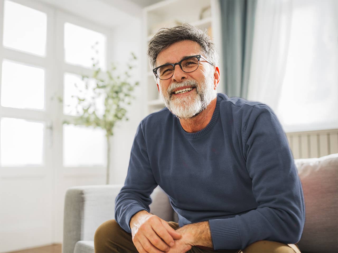 Older white man with a grey beard and glasses sits in a sunny room