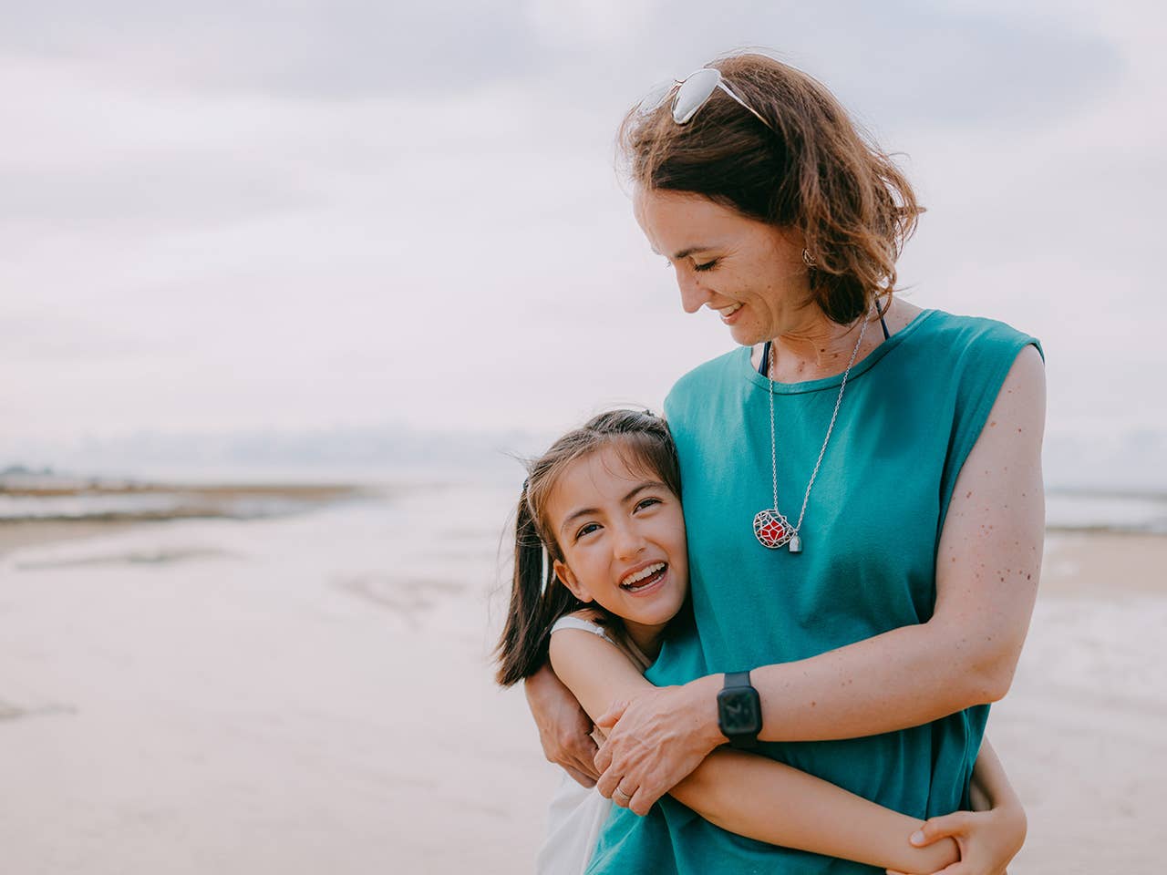 Smiling mother hugs her smiling daughter as they stand beside one another in the sunshine on a sandy beach