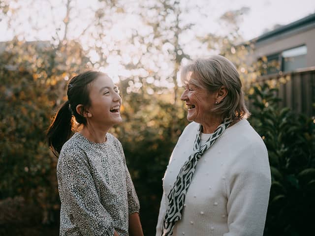 A teenage girl with black hair in a ponytail laughs while speaking to her grandmother outdoors as the sun sets behind trees