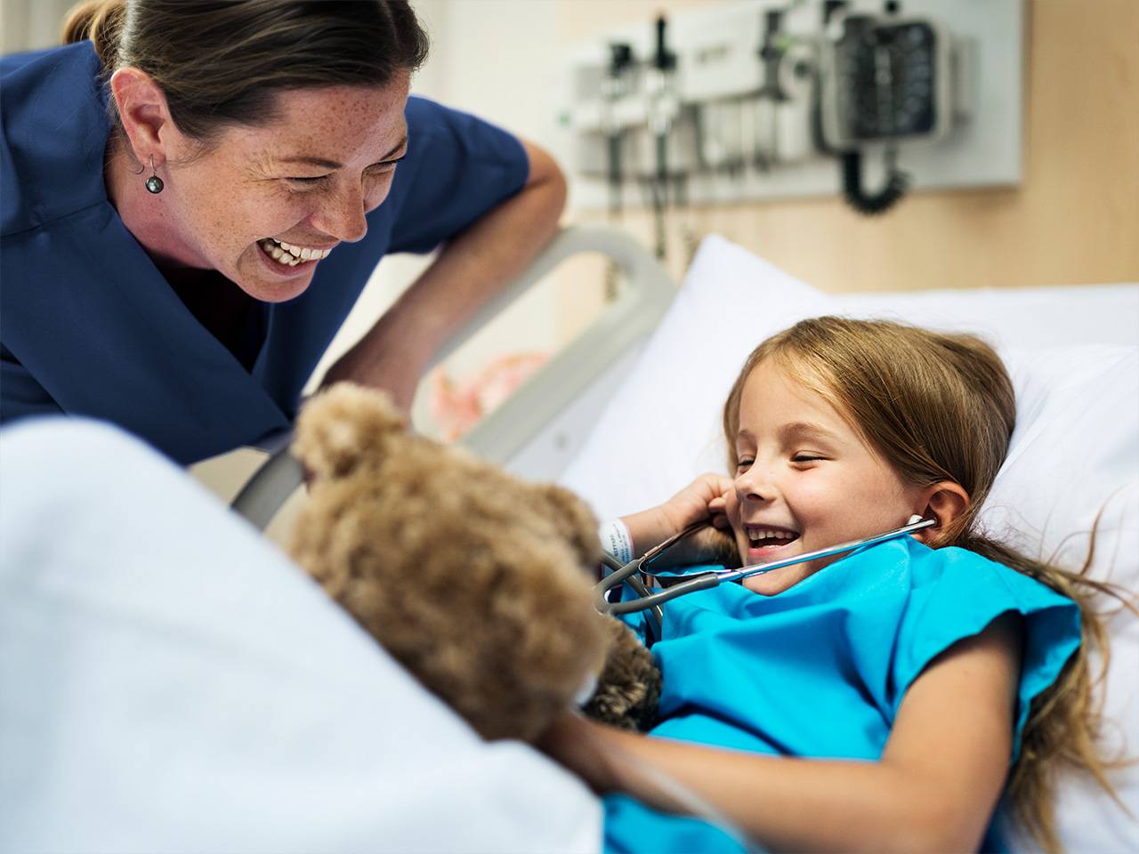 Female medical professional smiling and watching a young female patient using a stethoscope on her toy bear while lying in a hospital bed