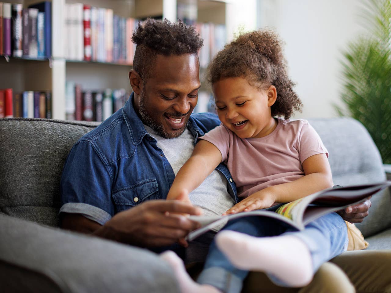 Smiling Black man sits with his young daughter on the sofa, reading a book together
