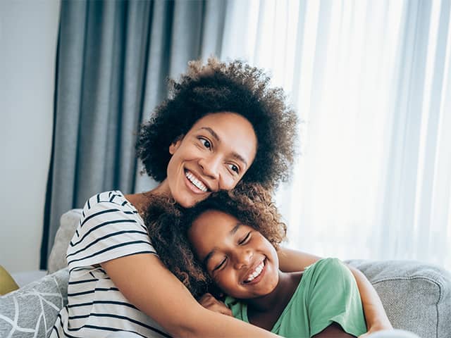 Smiling Black woman wearing a white shirt with navy blue stripes sits on a couch and hugs her smiling daughter