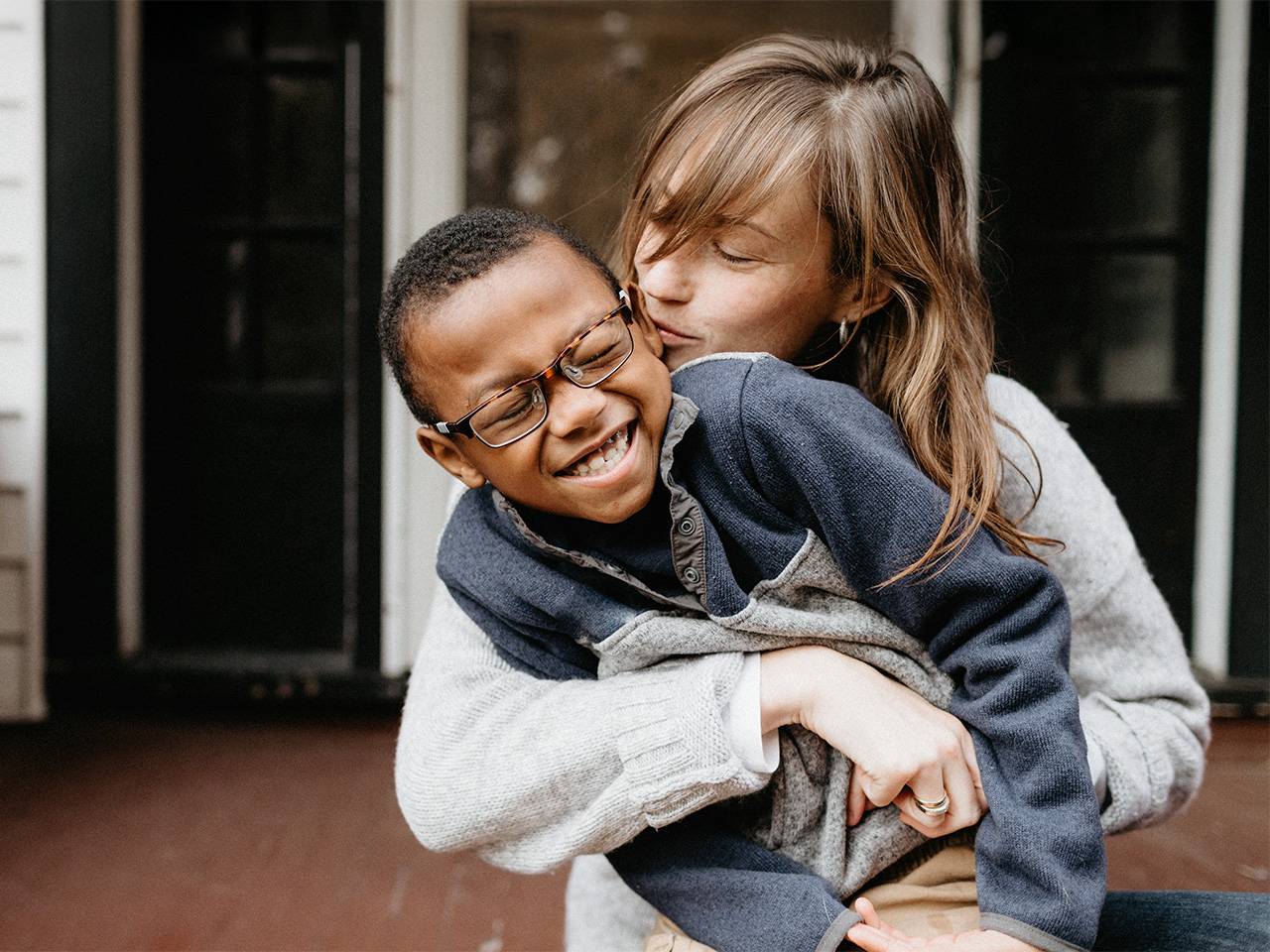 A mother playfully wraps her arms around her son and kisses his cheek as he laughs and leans towards his side