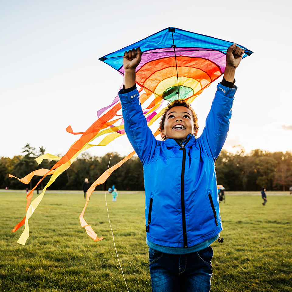 African American boy in a blue coat holds rainbow colored kite above his head