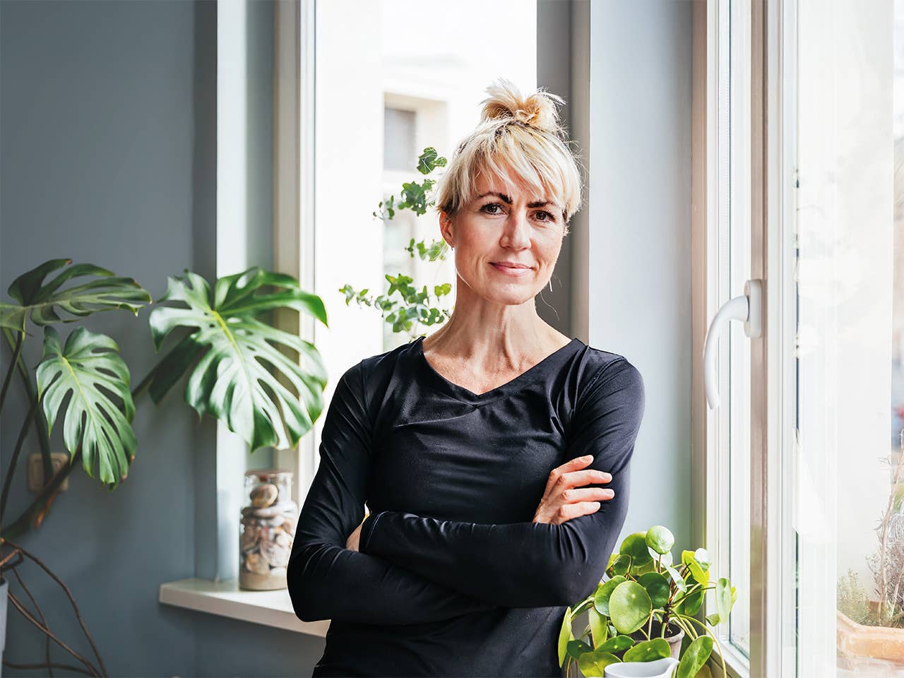 Middle aged white woman wearing a black shirt stands in a sunny room, crossing her arms over her torso