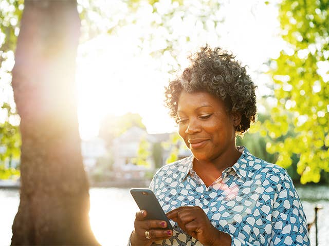 Black woman in a light blue shirt stands beneath a tree in the sunlight, looking at her phone
