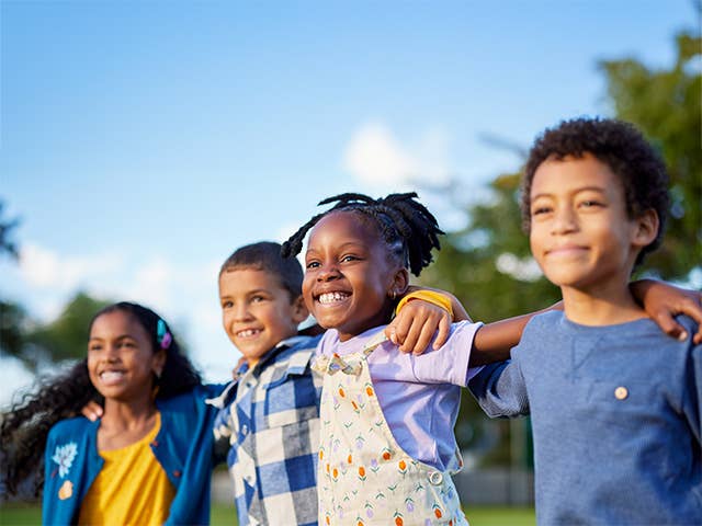Group of four smiling boys and girls link arms across one another's shoulders outdoors in the sunshine