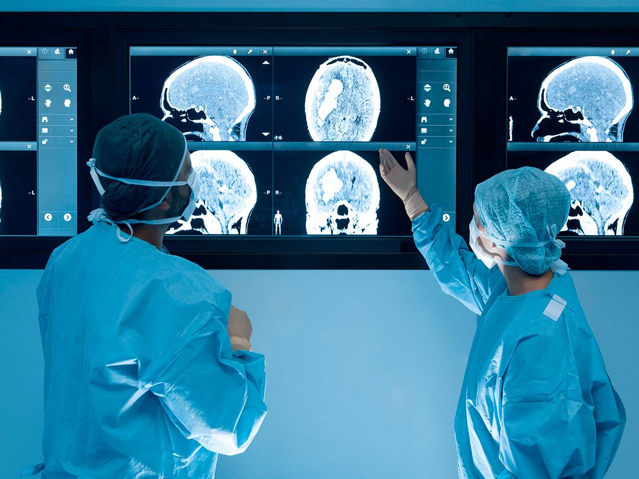Two surgical team members review brain scan images on large medical display screens.