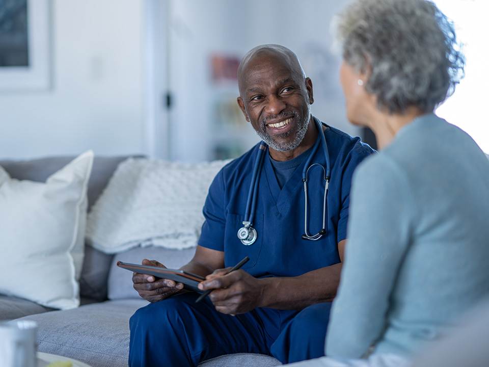 Smiling Corewell Health home based primary care provider wearing blue scrubs speaks to an elderly patient as they sit next to each other on a sofa