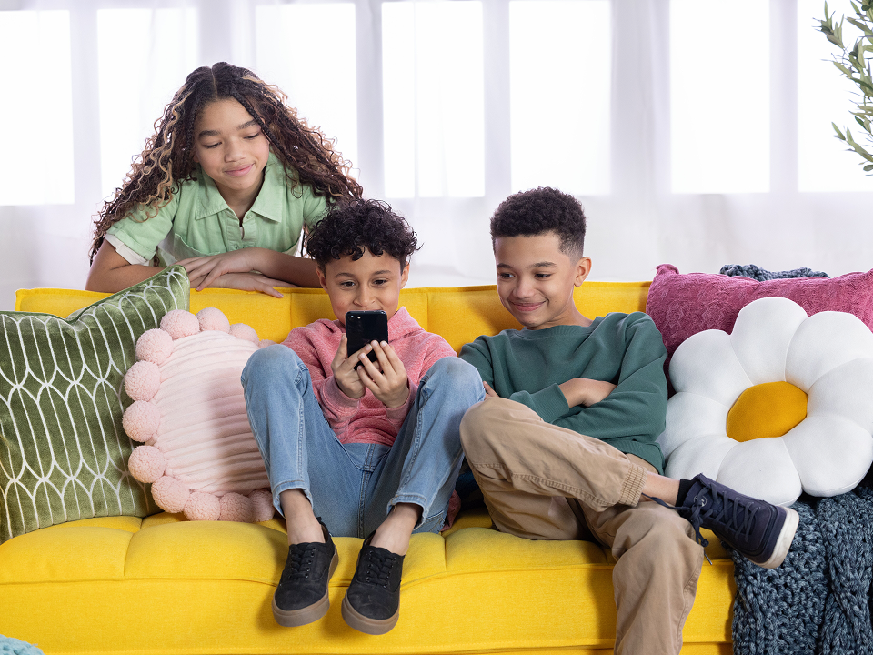 Three children sit on a couch, surrounded by colorful pillows, smiling and enjoying each other's company.