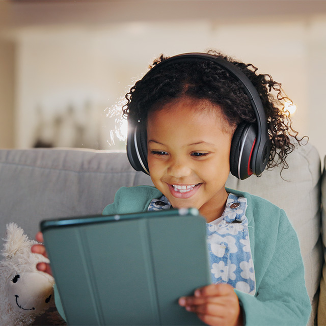 A young Black girl with curly brown hair smiles and looks at a tablet device while wearing headphones