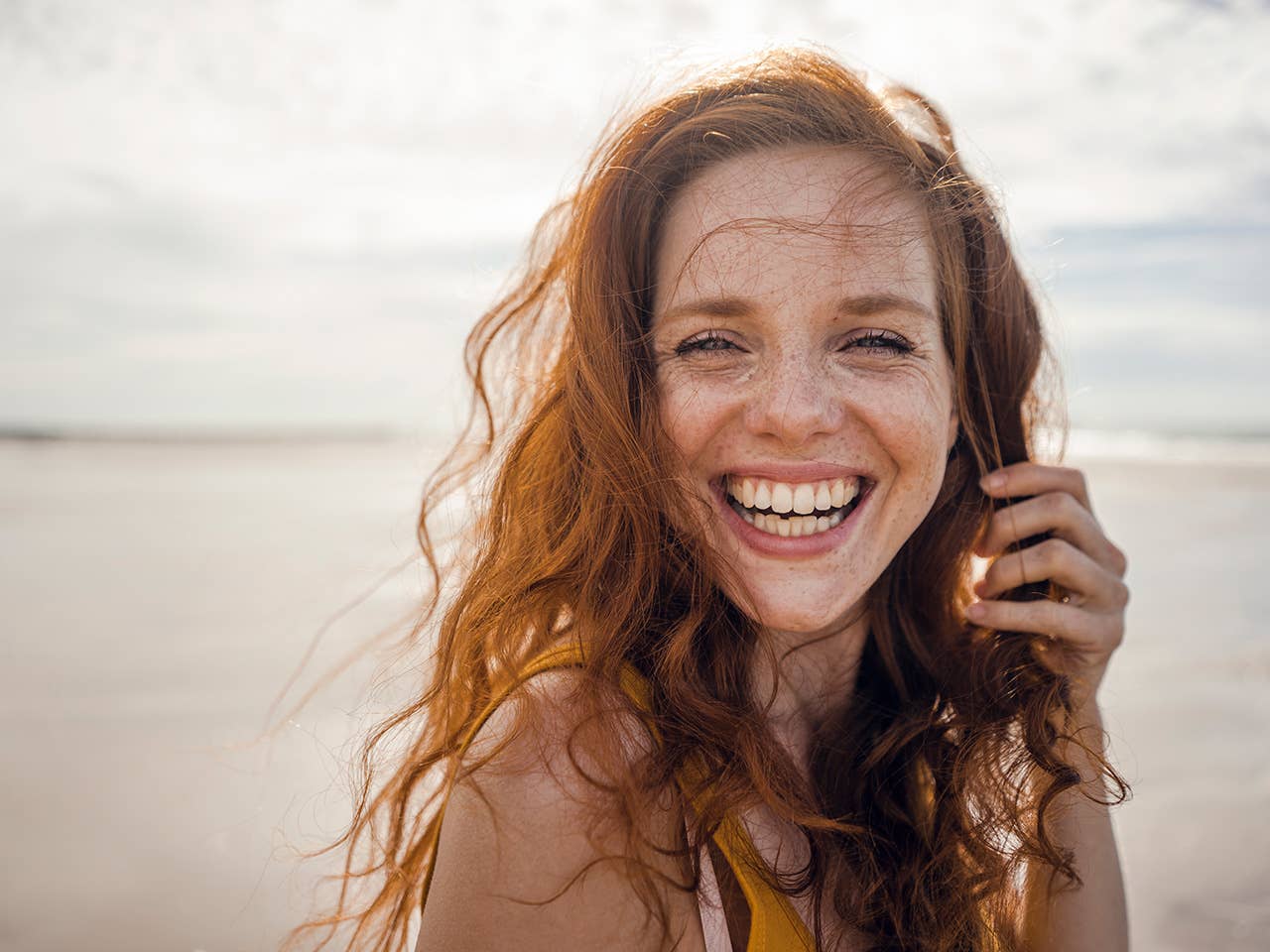 Smiling White woman with red hair and freckles stands outdoors along the beach