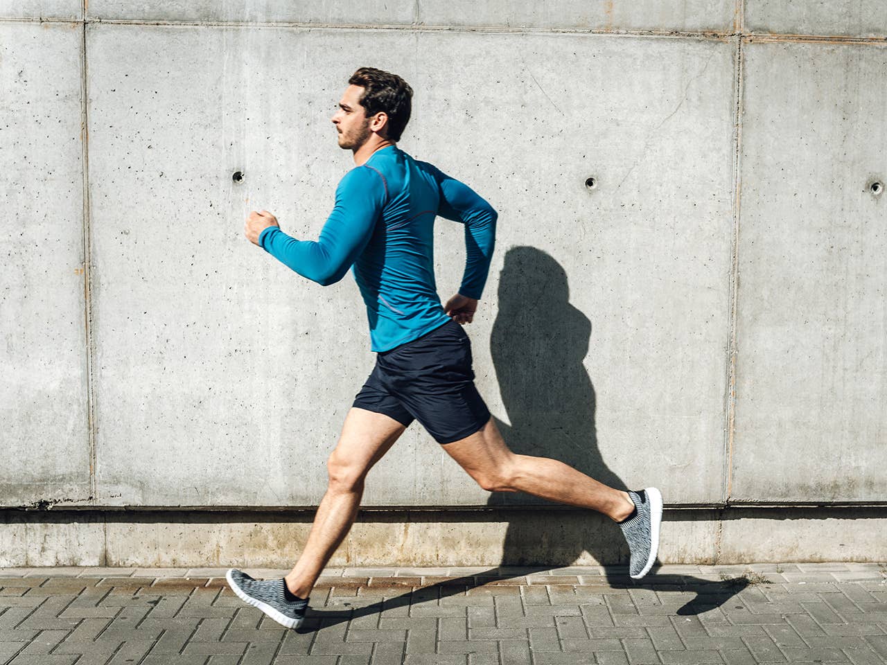 Young white man in black shorts and a blue shirt jogs on the sidewalk in sunshine