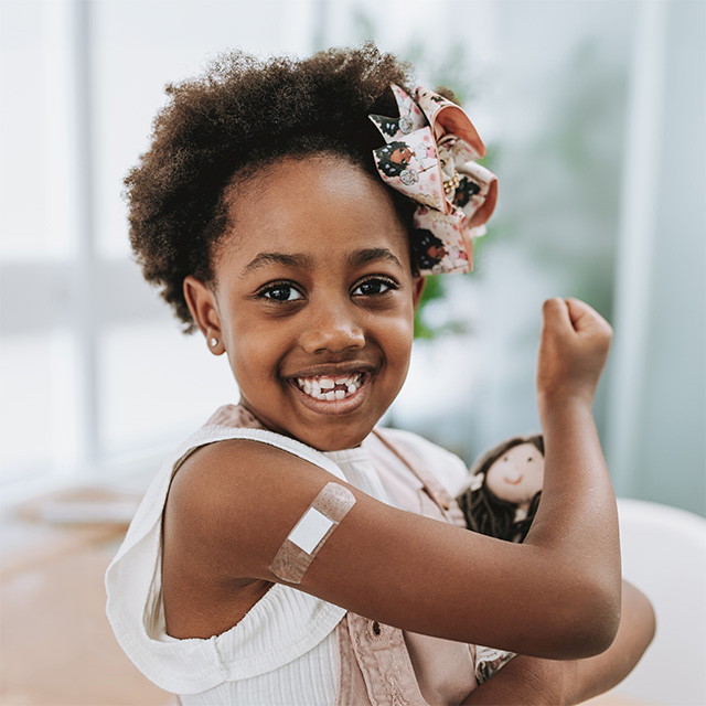 Child showing a bandage after a flu vaccine, holding a doll and flexing an arm in a healthcare setting.