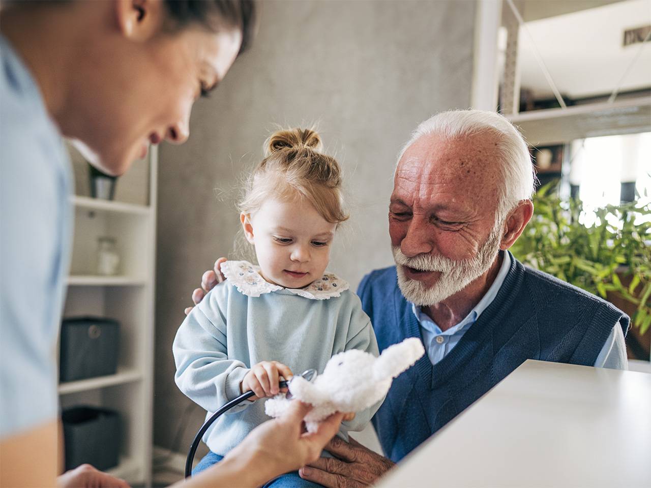 Child using a stethoscope on a stuffed animal while seated with an older adult as a healthcare worker assists.