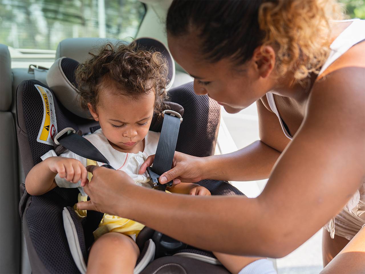 A young mother leans over her toddler while she straps her into a car seat