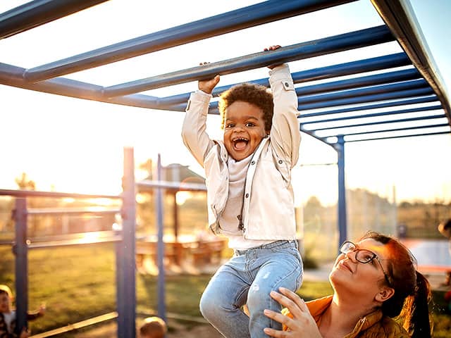 Smiling young Black boy wearing jeans and a white jacket hangs from monkey bars on a playground