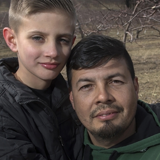 Preteen boy stands next to his father in front of small trees in the autumn