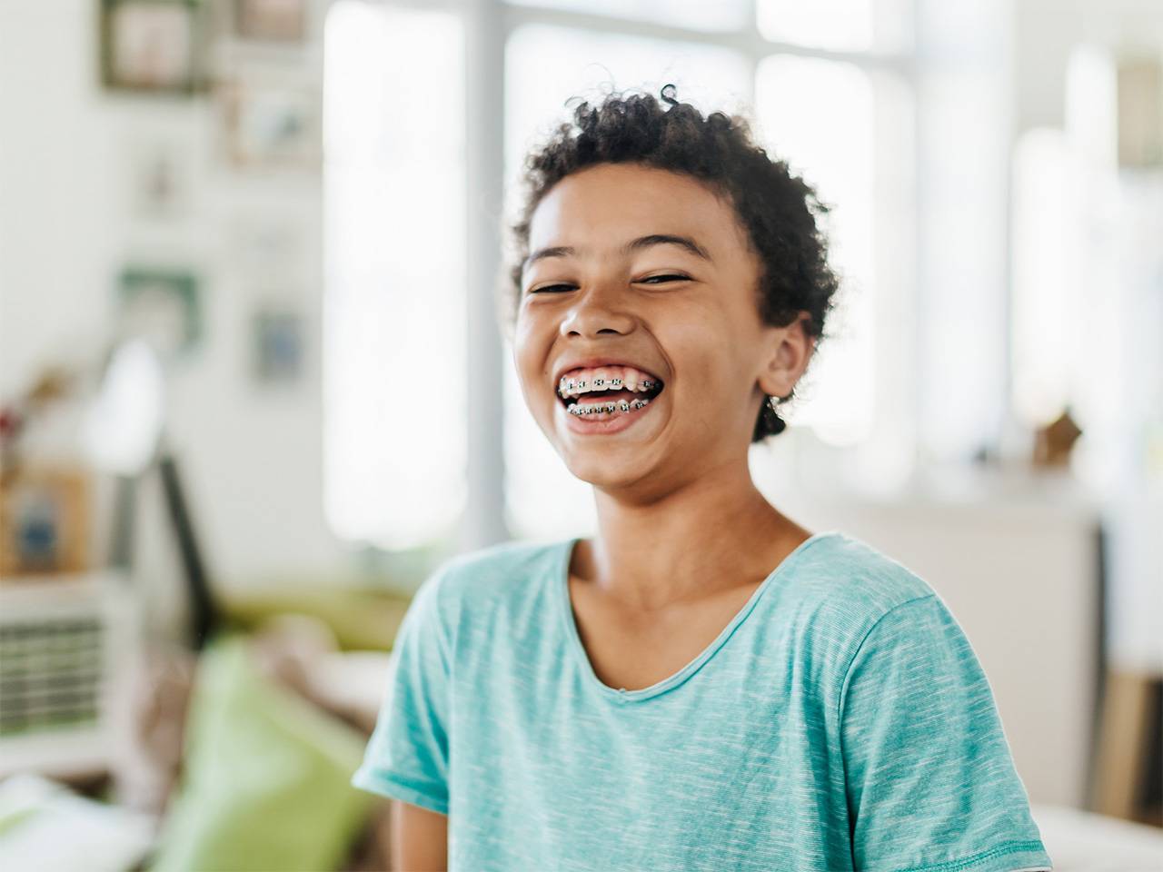 Girl with short curly brown hair and braces on her teeth smiles and laughs inside a sunny room