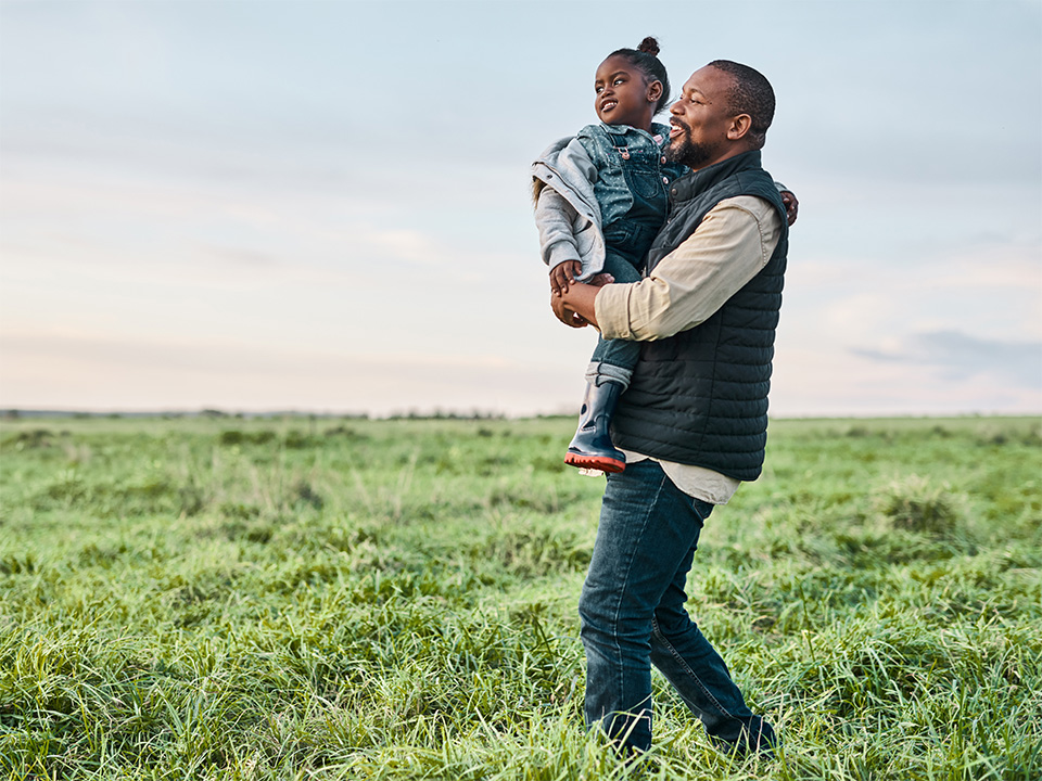A man holds a child in a grassy field, both smiling and enjoying a sunny day together.