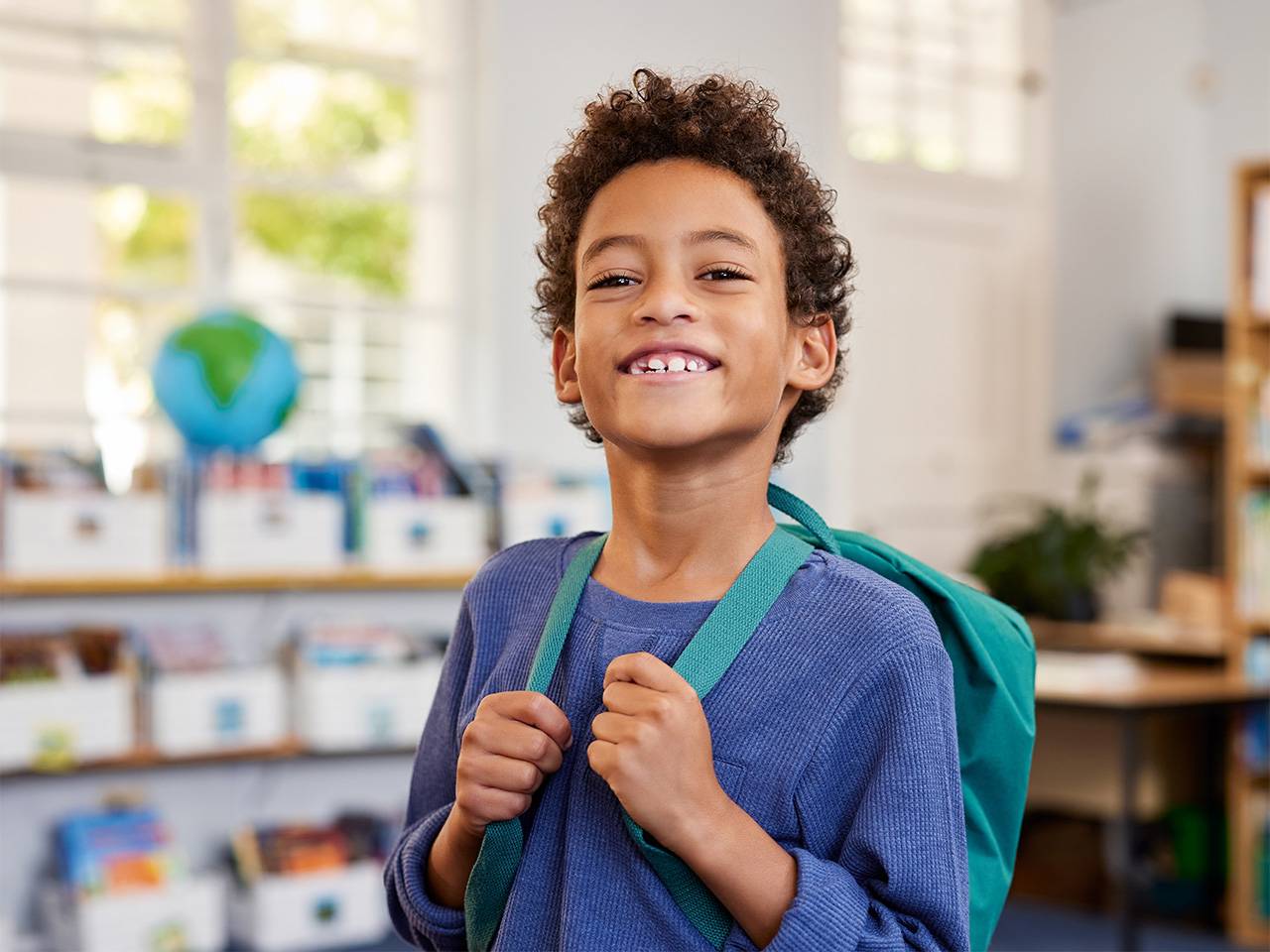 Smiling Black boy wearing a blue shirt stands with his backpack inside a classroom