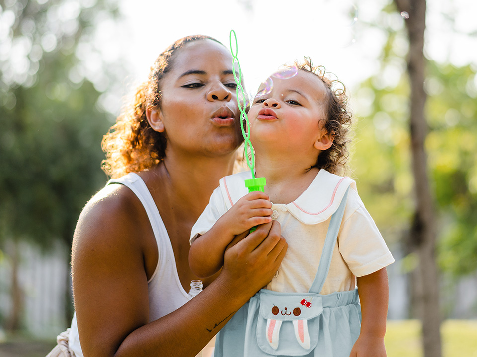 Young woman and her toddler daughter blow bubbles from a bubble wand in the sunshine outdoors