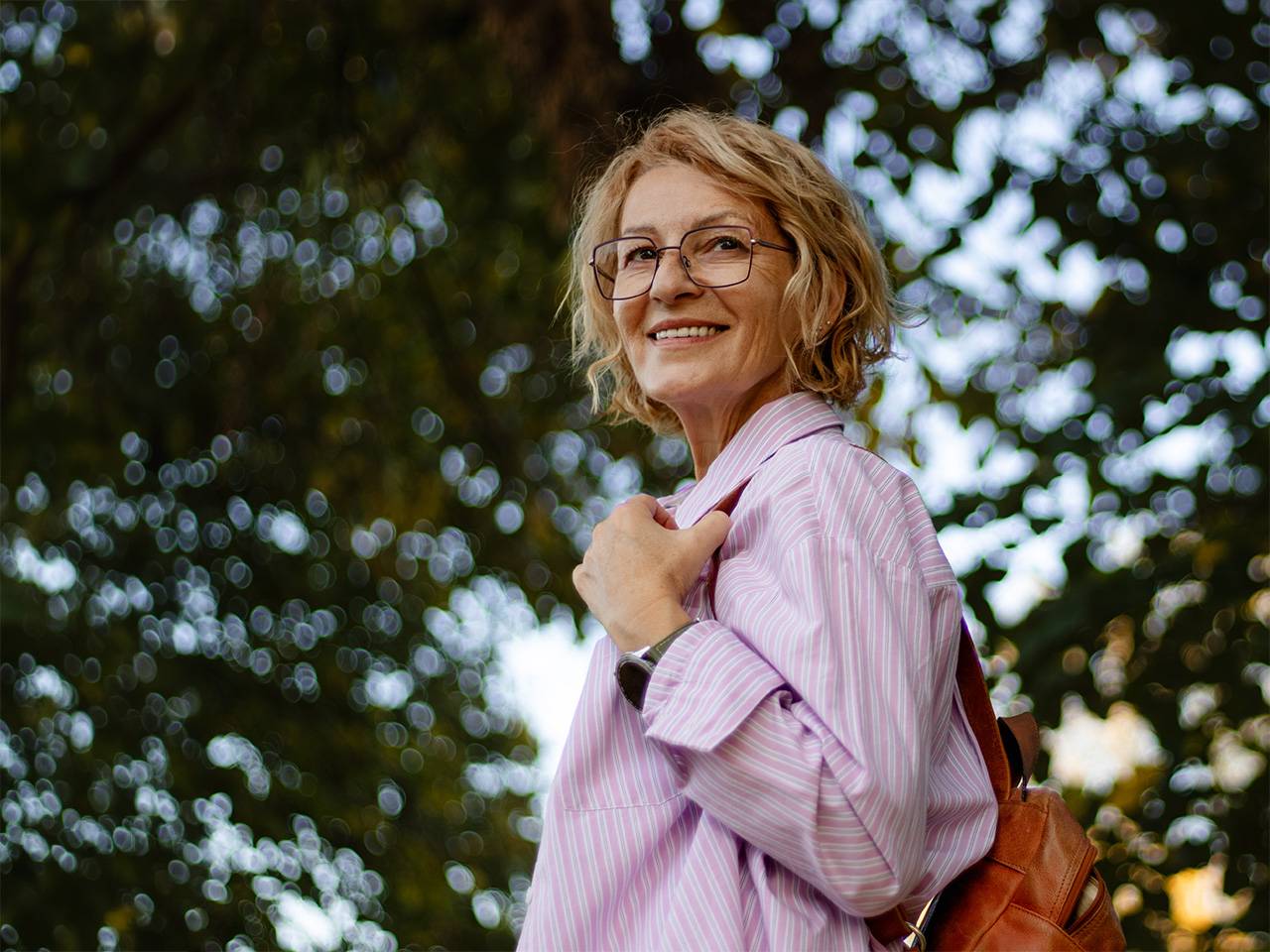 Smiling older white woman wearing glasses looks over her shoulder while in the shade of a tree outdoors