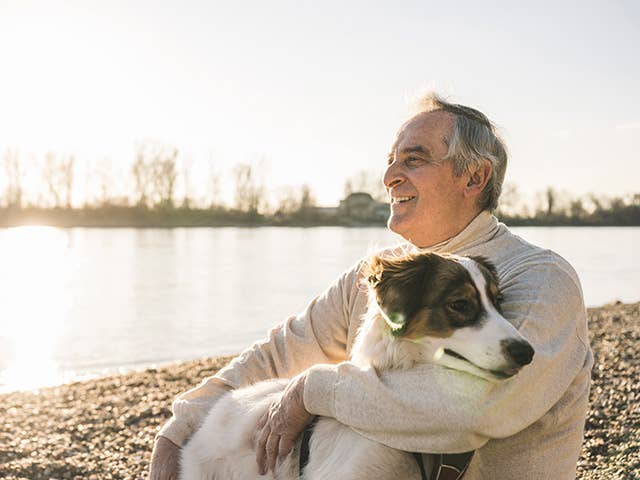 Older white man wearing a white sweater sits with a dog next to a small lake