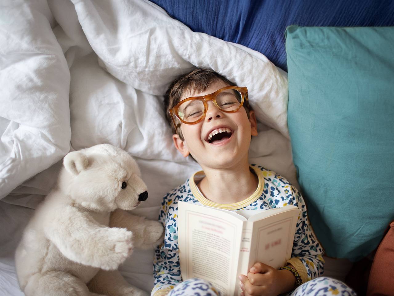 Boy wearing pajamas and glasses lies in bed and laughs while reading a book