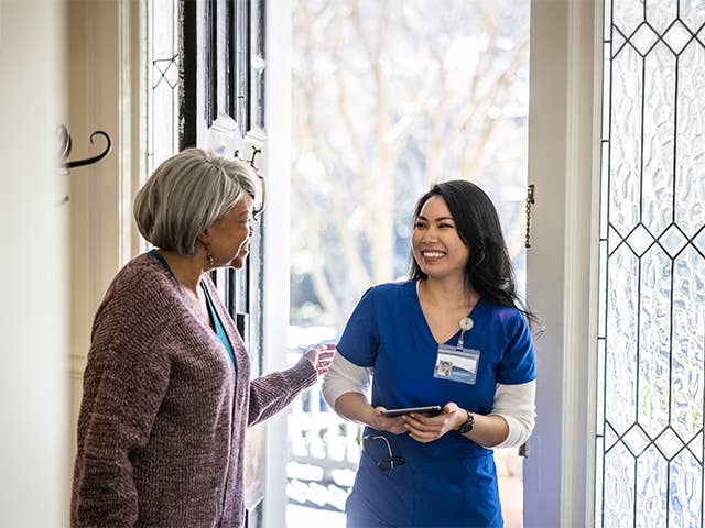 Smiling Corewell Health home based primary care provider wearing blue scrubs speaks with an elderly woman inside her front door