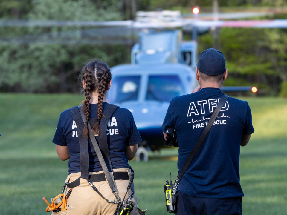 A male first responder wearing a navy blue shirt and ballcap stands next to a female first responder wearing a navy blue shirt in a grassy field, as an Aero Med helicopter sits in the background