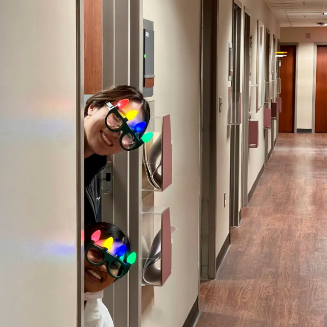 Two female residents wearing Christmas themed headbands peeking around the corner of a door into the hallway at the hospital