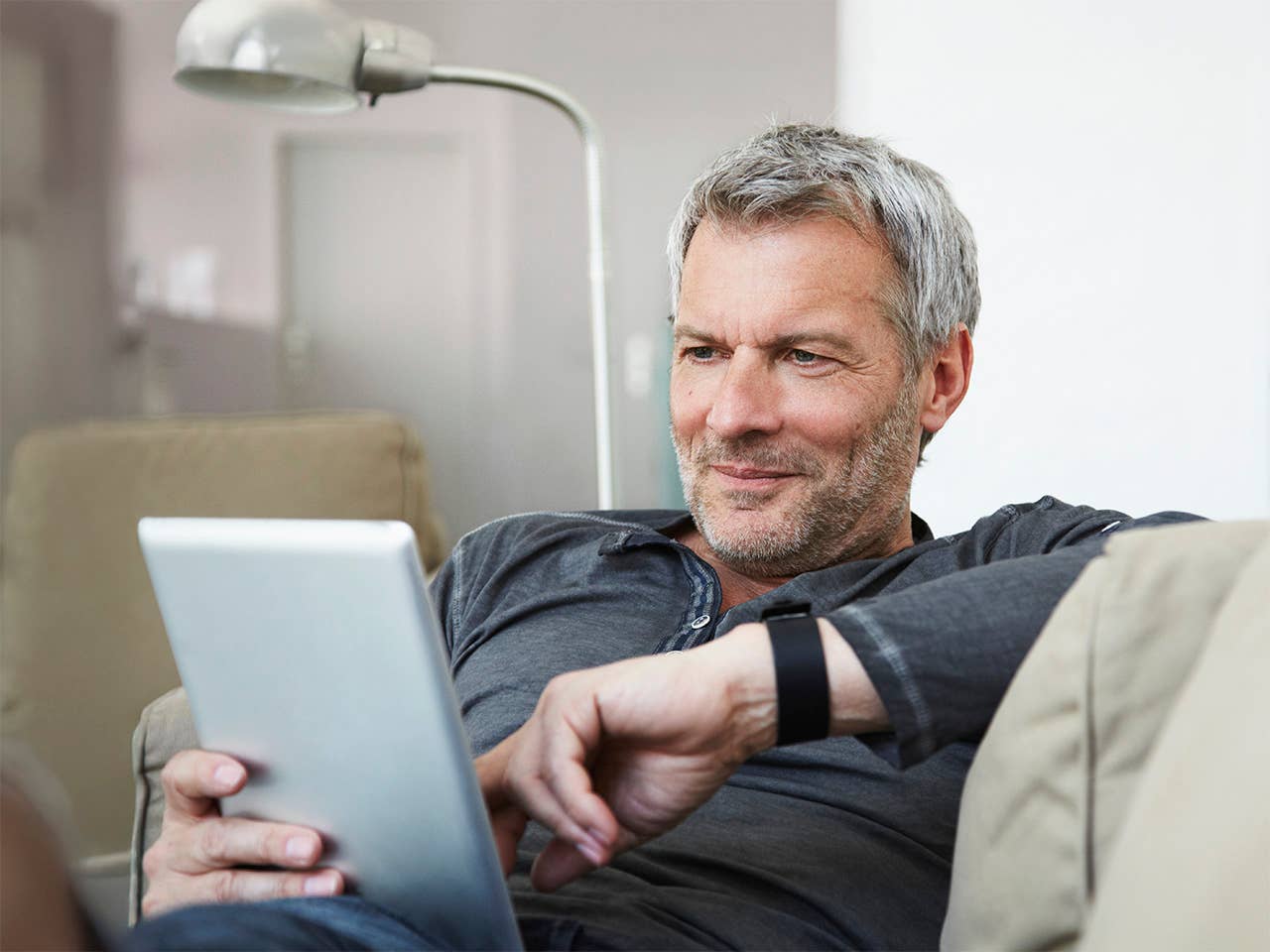 Grey-haired white man sits on a couch looking at a tablet he is holding
