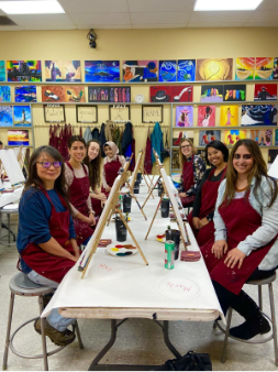 A group of women gathered around a table, each holding paint brushes and engaged in a creative painting activity.