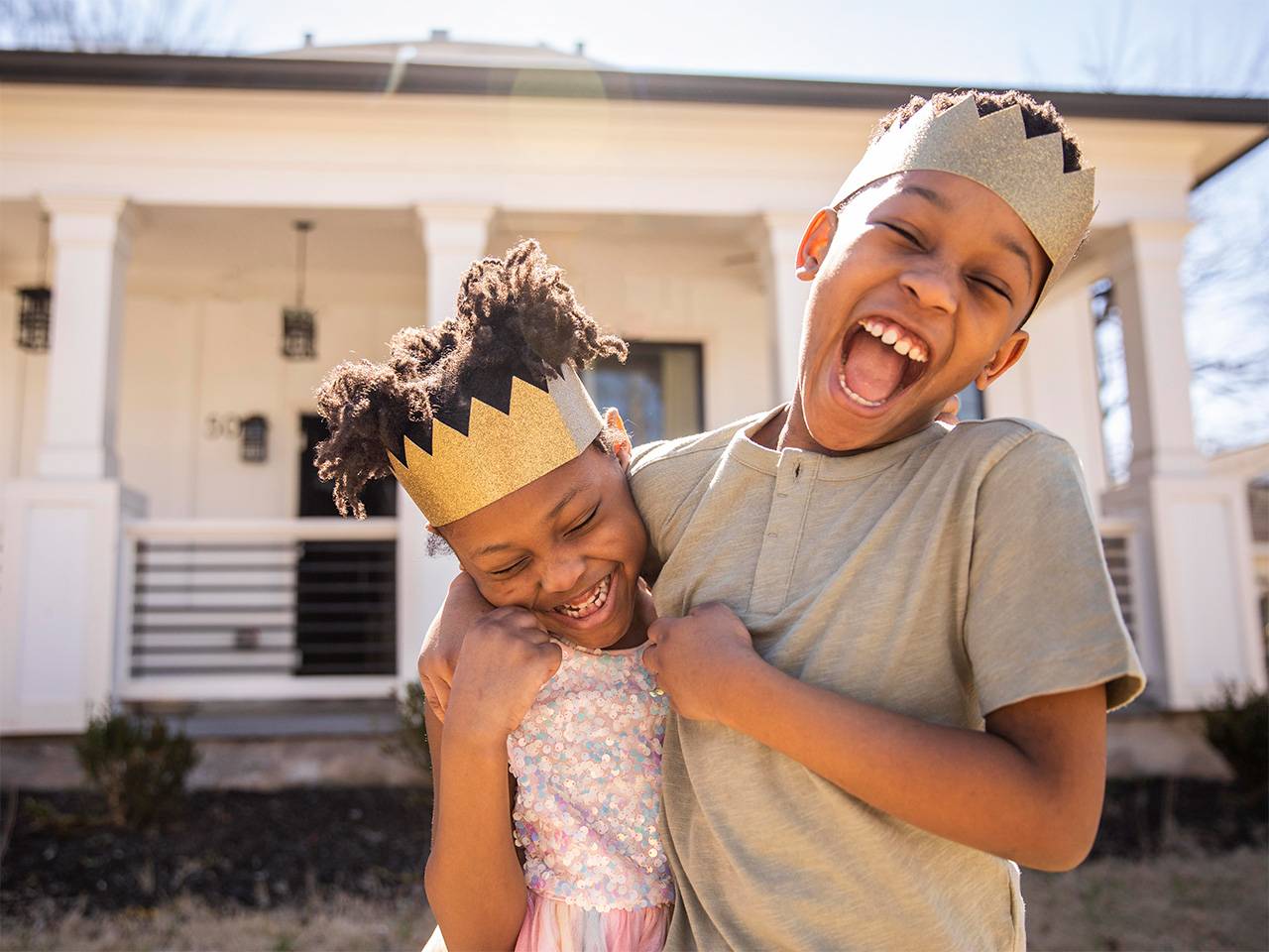 Smiling Black boy wearing a toy crown wraps his arm around his smiling sister as they play outdoors