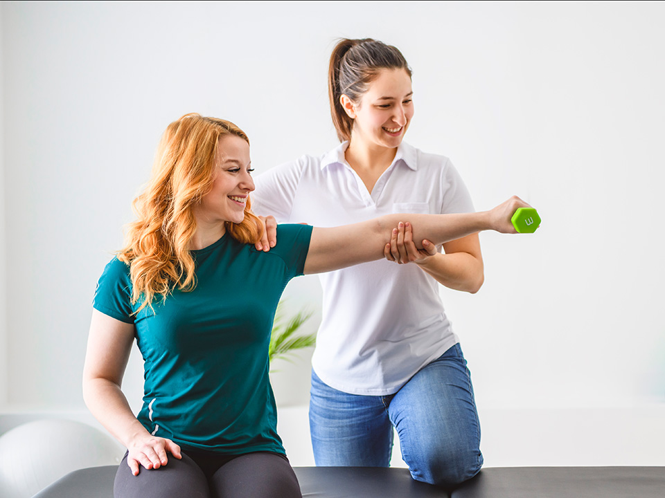 Therapist helps a smiling woman hold out her arm while holding a small dumbell