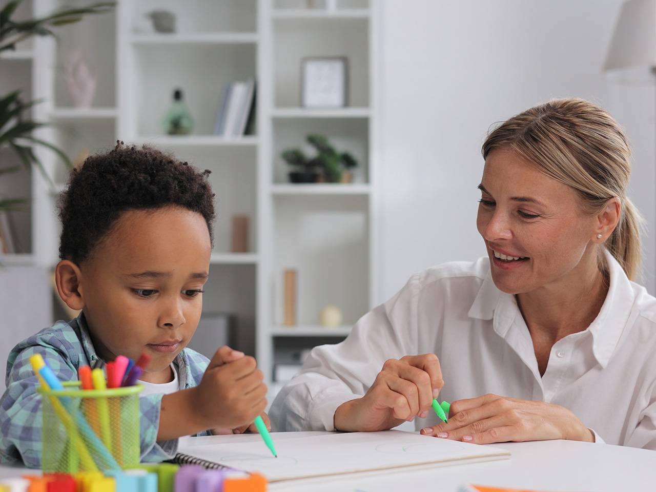 Smiling woman wearing a white blouse sits next to a young Black boy at a table as they color with markers