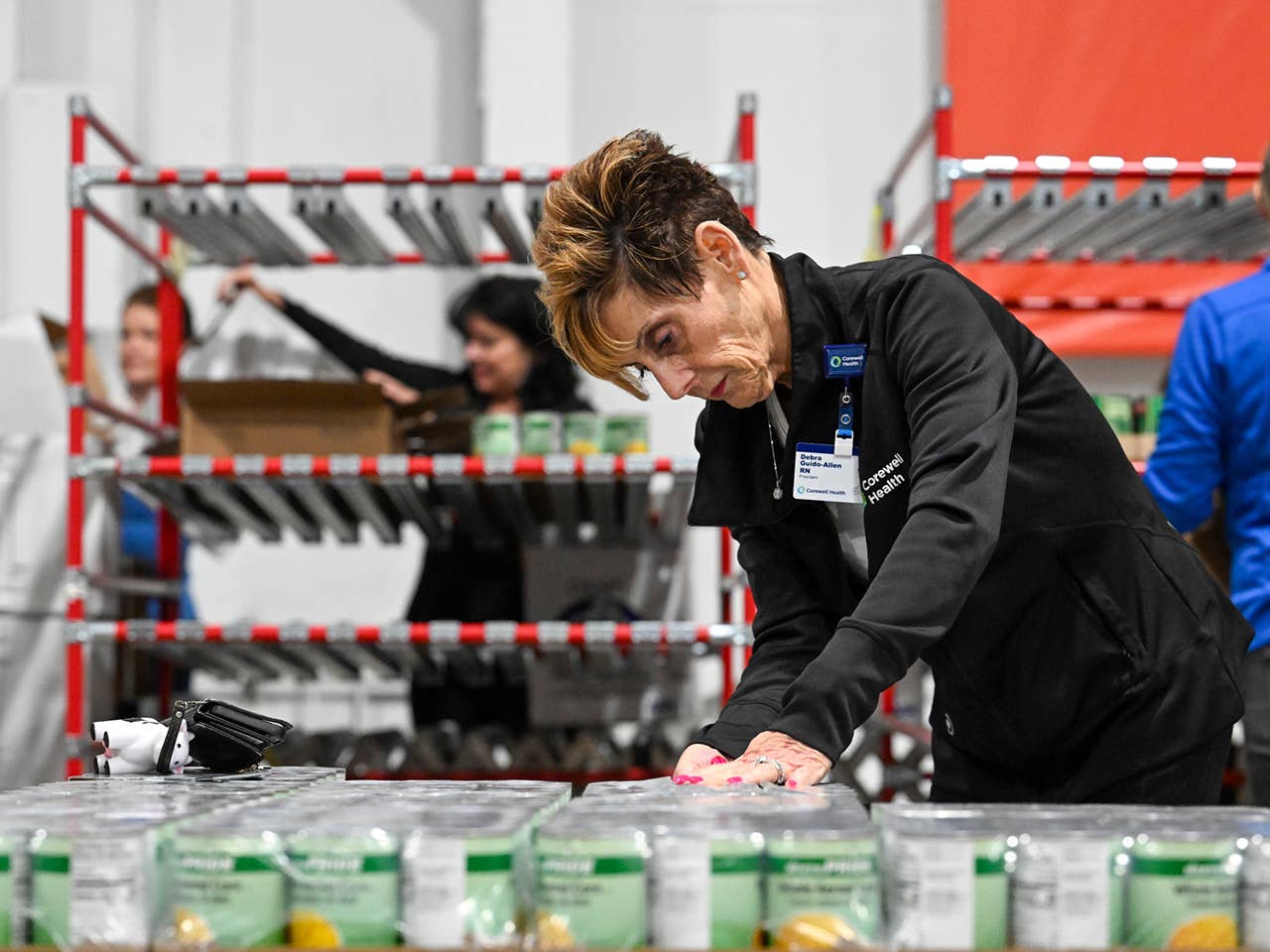 A woman stands in a warehouse surrounded by stacked cans and boxes, organizing supplies for distribution.