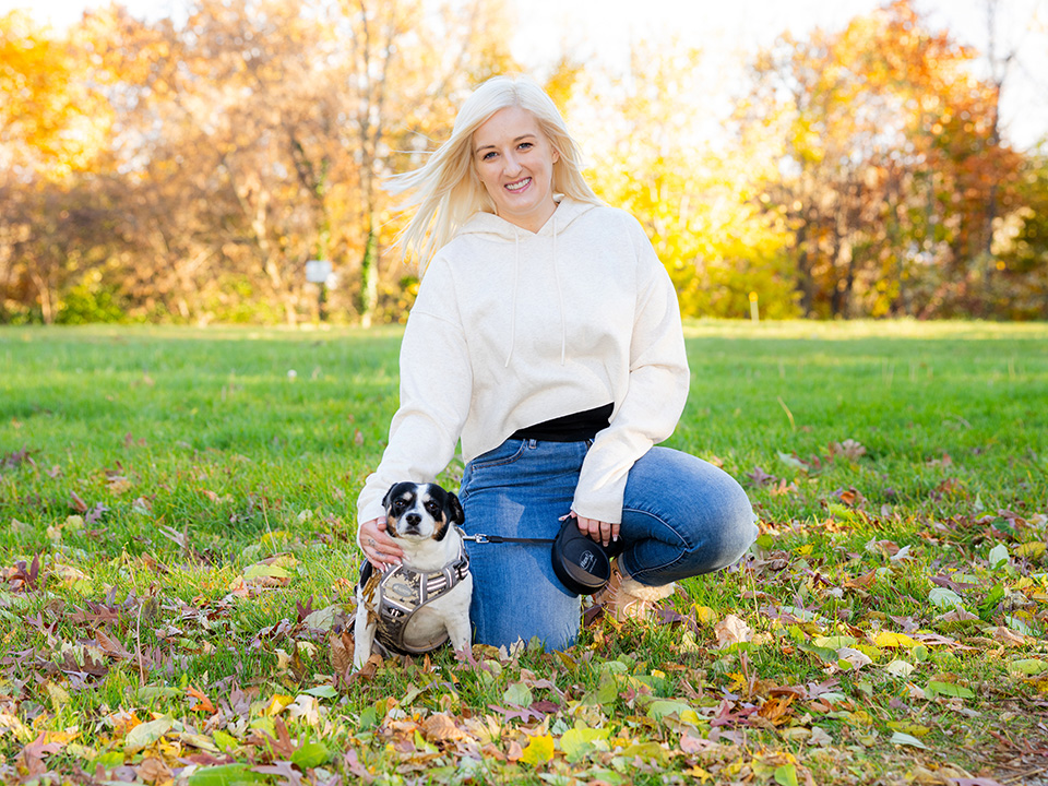 Young white woman kneels down in a grassy field next to a dog