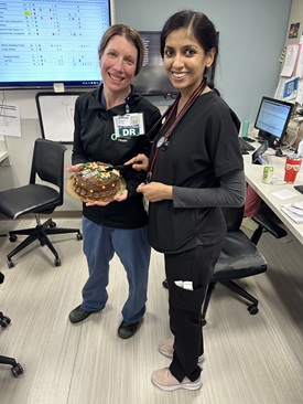 In an office setting, two women in scrubs joyfully hold a cake, symbolizing the commitment to wellness among medical fellows.