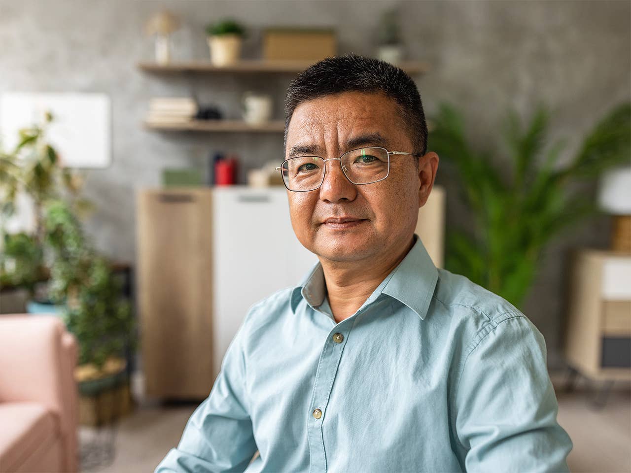Middle aged Asian man in a blue shirt and wearing glasses sits in a room with plants behind him