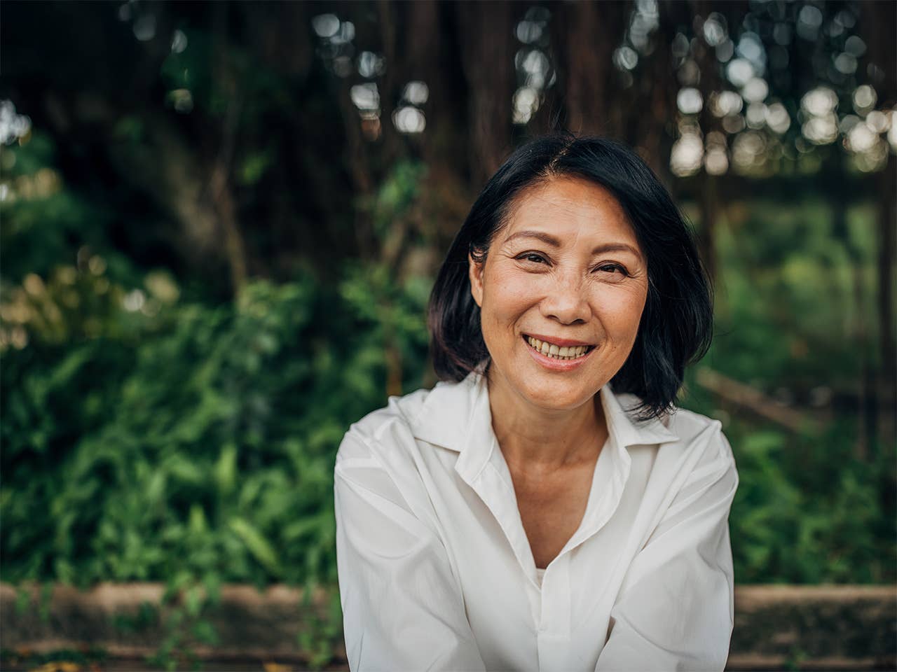 Asian woman in a white blouse sitting on a  bench in front of bushes