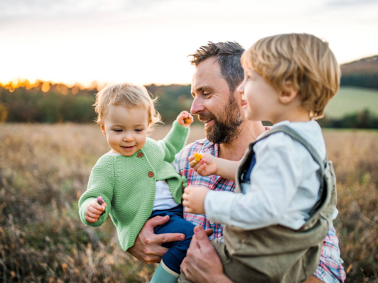 Middle aged man holds two toddlers as he stands in a field in the evening