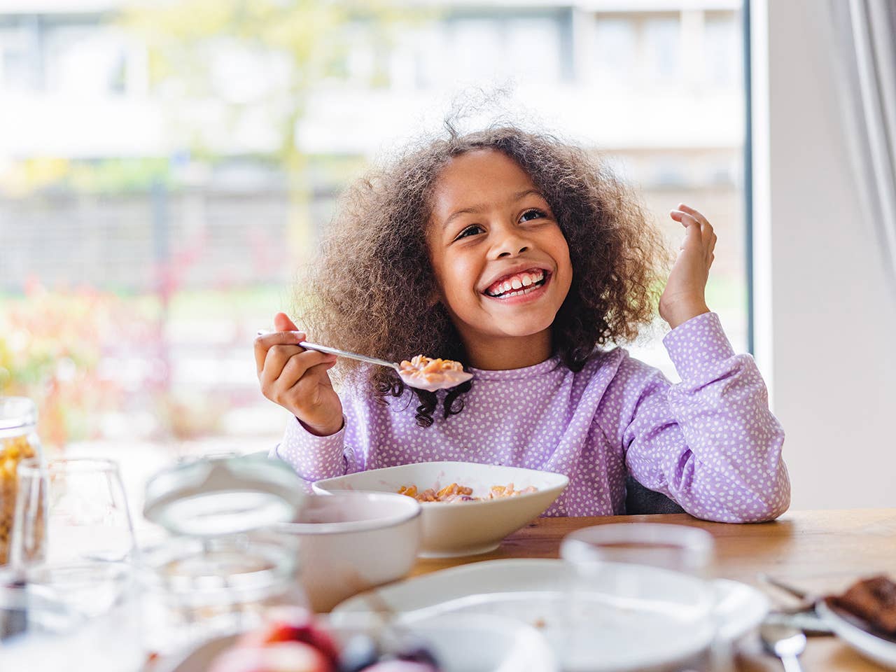 Smiling Black girl with curly brown hair sits at a table with a spoon in her hand as she eats from a bowl of cereal